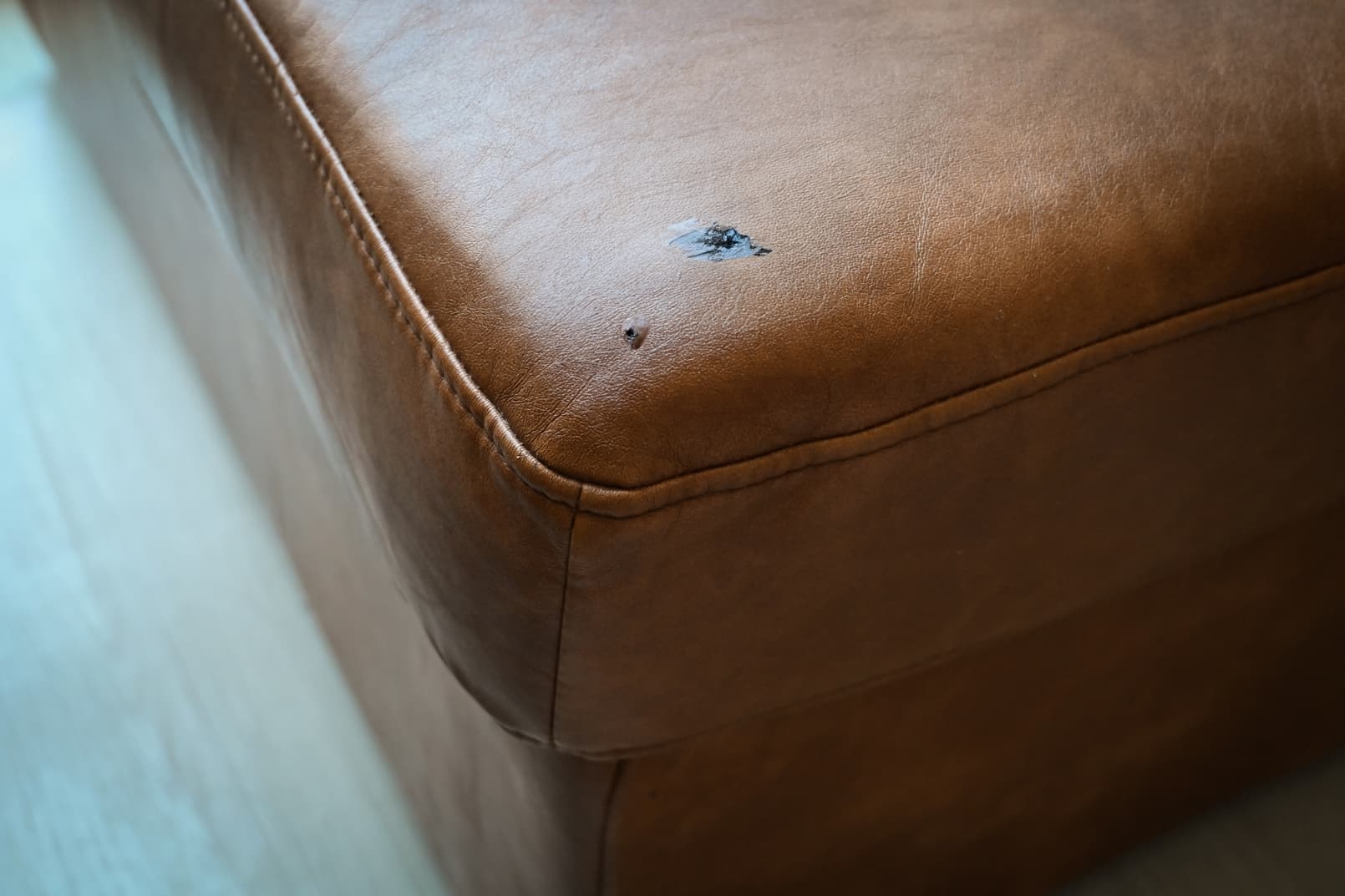 Een close-up van een bruine leren bank met een zichtbare scheur en schaafplek op de bovenste hoek van het kussen.