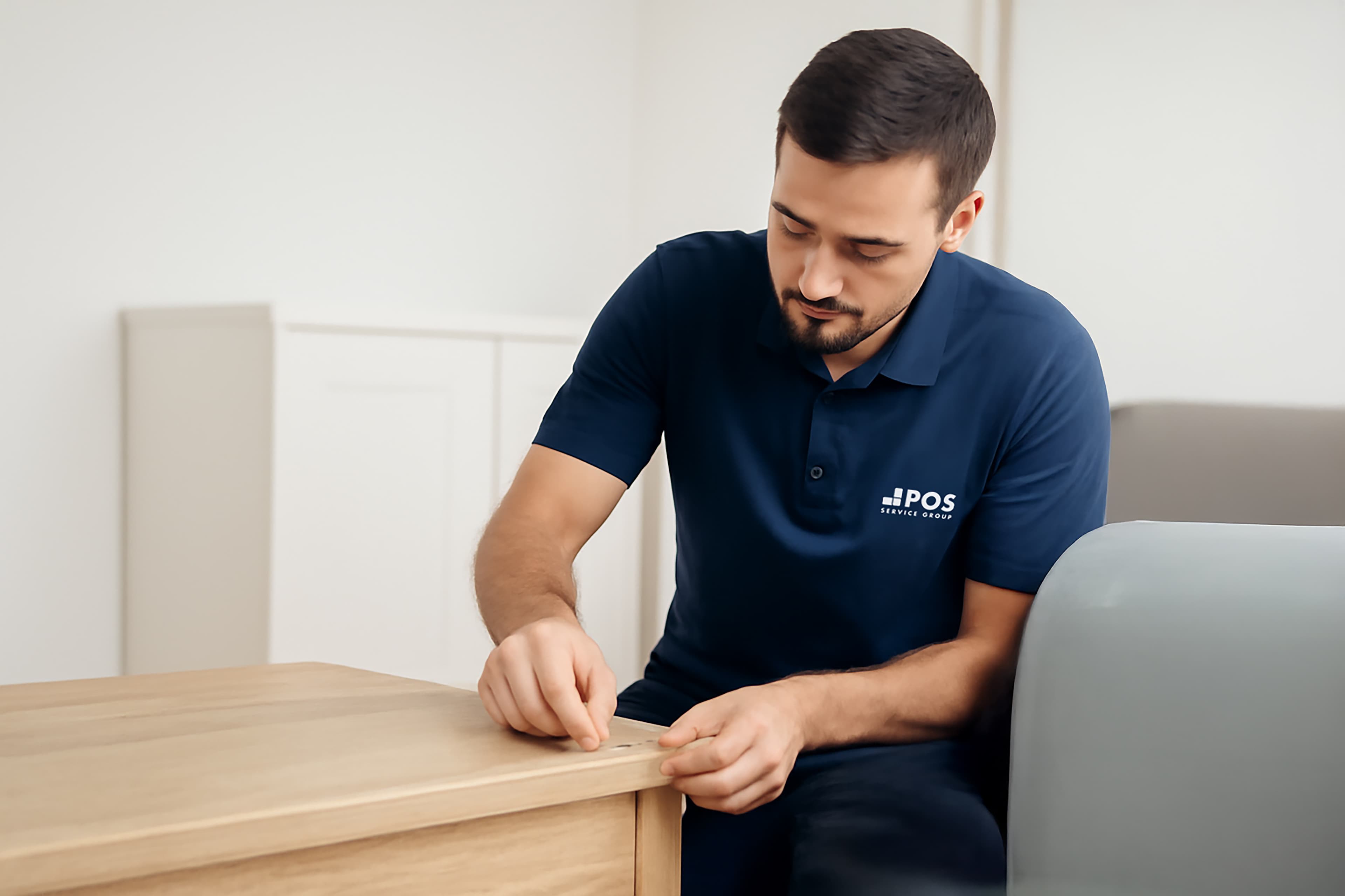 A man in a navy blue uniform assembles a wooden piece of furniture, focusing intently as he screws a fastener into the wood.