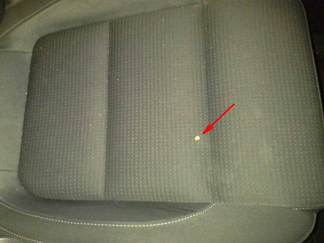 A small round object is on a dark, textured car seat. A red arrow points directly at the object, highlighting its location.