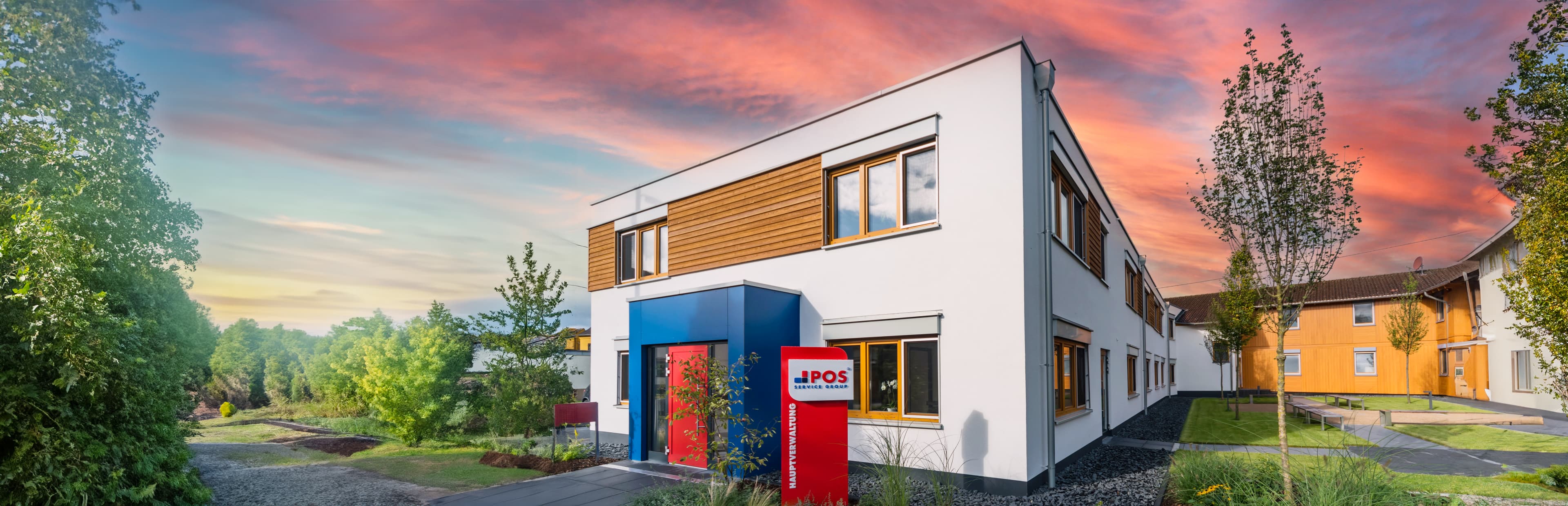 A modern, two-story white building with wooden accents and large windows stands under a colorful sunset sky. A red sign reading “IPOS” is near the entrance, surrounded by trees and greenery.