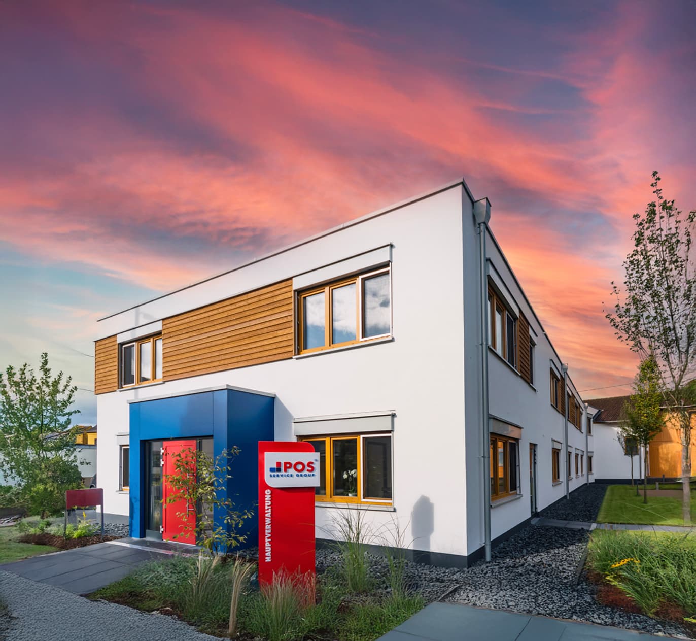 Modern two-story white building with wooden accents and large windows, featuring a bold red and blue entrance. A red POS sign stands out front. The sky is vibrant with pink and orange clouds at sunset.